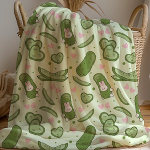 May include: A light yellow blanket featuring a repeating pattern of green pickles, pink hearts, and cartoon bunnies dressed as pickles. The blanket is draped over a wicker basket.