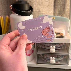 May include: A stack of lavender-colored cards with the text "I'M HAPPY" and a cartoon dog illustration. The cards feature a moon, stars, and clouds design. The cards are held in a hand.