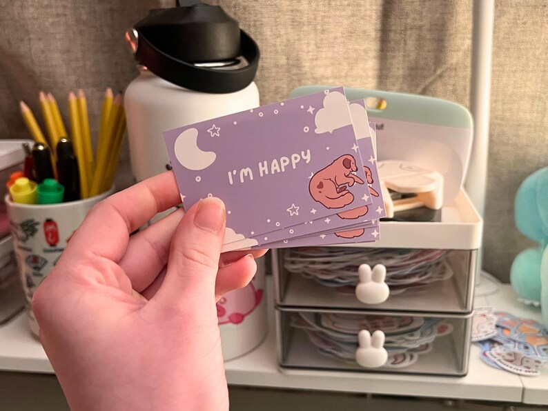 May include: A stack of lavender-colored cards with the text "I'M HAPPY" and a cartoon dog illustration. The cards feature a moon, stars, and clouds design. The cards are held in a hand.