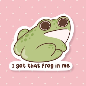 May include: A sticker featuring a cartoon frog in shades of green, with large brown eyes and a white belly. The text "I got that frog in me" is below the frog. The background is pink with white hearts.