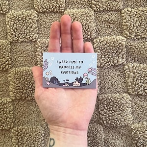 Puede incluir: Una peque&ntilde;a tarjeta rectangular con el texto "I NEED TIME TO PROCESS MY EMOTIONS" se sostiene en una mano. La tarjeta presenta una escena submarina con corales, medusas y otras criaturas marinas en tonos de azul, gris y rosa.