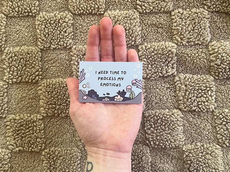 Puede incluir: Una peque&ntilde;a tarjeta rectangular con el texto "I NEED TIME TO PROCESS MY EMOTIONS" se sostiene en una mano. La tarjeta presenta una escena submarina con corales, medusas y otras criaturas marinas en tonos de azul, gris y rosa.