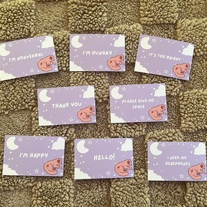 May include: A collection of lavender-colored cards with various phrases and illustrations. Each card features a sleeping creature, a crescent moon, clouds, and stars. Phrases include "I'm Nonverbal," "I'm Hungry," and "Hello!"
