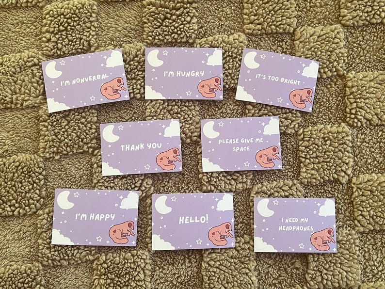 May include: A collection of lavender-colored cards with various phrases and illustrations. Each card features a sleeping creature, a crescent moon, clouds, and stars. Phrases include "I'm Nonverbal," "I'm Hungry," and "Hello!"
