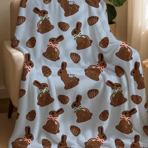 May include: A light blue blanket featuring a repeating pattern of chocolate Easter bunnies and chocolate eggs. The bunnies have pink or green bows. The blanket is draped over a beige armchair.