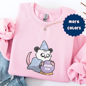 Puede incluir: Una sudadera rosa claro con un dibujo de un opossum de dibujos animados con un sombrero y capa de mago, sosteniendo una bola de cristal con las palabras "dump him". Un círculo azul marino con las palabras "more colors" está en la esquina superior derecha.