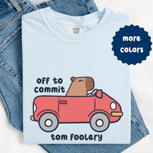 May include: Light blue t-shirt with a cartoon capybara driving a red car. Text on the shirt reads "off to commit tom foolery." A navy blue circle with the words "more colors" is on the upper right.