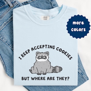 May include: Light blue t-shirt with a raccoon graphic and the text "I keep accepting cookies but where are they?" The shirt is folded on top of a pair of blue jeans. A navy blue circle with the words "more colors" is in the upper right corner.