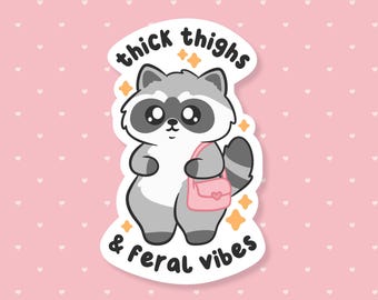 May include: A sticker featuring a cartoon raccoon with large eyes and a pink purse. The text "thick thighs & feral vibes" is written above and below the raccoon in a playful font. The background is pink with white hearts.