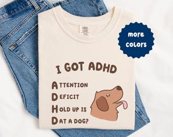 Puede incluir: Camiseta color crema con el texto "I GOT ADHD" y un desglose de acrónimos. Un perro de dibujos animados con la lengua fuera está a la derecha. La camiseta está colocada encima de unos vaqueros azules. Un círculo azul marino con las palabras "more colors" está arriba a la derecha.