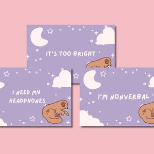 May include: Three lavender-colored cards with illustrations of a sleeping dog, moon, clouds, and stars. Text on the cards reads: "It's too bright," "I need my headphones," and "I'm nonverbal."