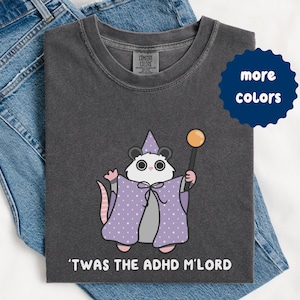 May include: Dark gray t-shirt featuring a cartoon opossum dressed as a wizard, wearing a purple cape with white polka dots and a matching hat. The opossum holds a wand. Text at the bottom reads, "'TWAS THE ADHD M'LORD."