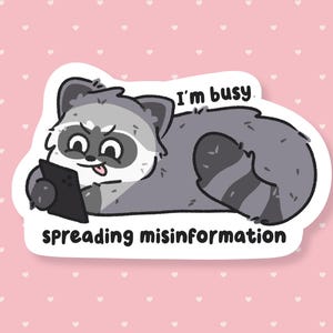 May include: A sticker featuring a cartoon raccoon lying down, holding a tablet. The raccoon is gray and white with a black mask and a tongue sticking out. Text on the sticker reads "I'm busy spreading misinformation."