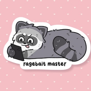 May include: A sticker featuring a cartoon raccoon lying down and holding a tablet. The raccoon is gray and white with a black mask and a tongue sticking out. The text "ragebait master" is below the raccoon.