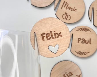May include: A collection of circular light-colored wooden drink markers featuring laser-engraved names including Felix, Johanna, Marie, Paul, and Mia. Each marker has a vertical slit for attachment to a glass rim and decorative icons.