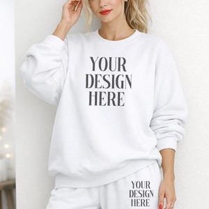 Op de afbeelding: Een witte sweatshirt en joggingbroek set. De sweatshirt en joggingbroek hebben de tekst "YOUR DESIGN HERE" in het zwart gedrukt. Het model leunt tegen een witte muur.