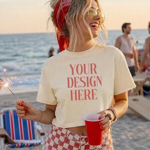 May include: A woman wearing a cream t-shirt with the text "YOUR DESIGN HERE" in red, and red and white checkered shorts. She holds a red cup and a sparkler on a beach. The scene is set on a sandy beach.