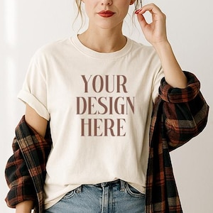May include: Cream-colored t-shirt with the text "YOUR DESIGN HERE" in brown lettering. The shirt is paired with light blue ripped jeans and a brown and black plaid flannel shirt. A clear plastic cup with a green straw is held in the hand.