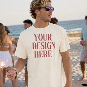 May include: Cream-colored t-shirt with the text "YOUR DESIGN HERE" in red. The person is wearing red and white checkered shorts and sunglasses. A red cup is held in the right hand. People are in the background on a sandy beach.