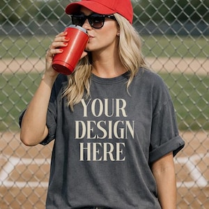 May include: A person wearing a red baseball cap, sunglasses, and a gray t-shirt with the text "YOUR DESIGN HERE." The person is holding a red tumbler. The background includes a chain-link fence and a baseball field.