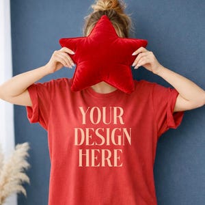 May include: A red t-shirt with the text "YOUR DESIGN HERE" in a light beige colour. A red star-shaped cushion covers the face. The shirt has rolled-up sleeves. The background is a dark blue wall.