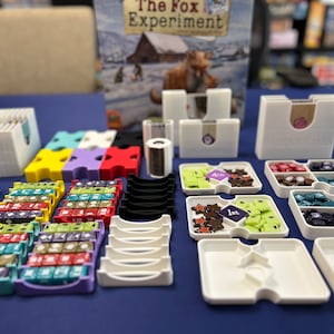 Puede incluir: Un juego de mesa llamado "The Fox Experiment" con piezas de juego coloridas, bandejas y una caja con una ilustración de un zorro. El juego incluye una variedad de componentes, incluyendo pequeños cubos, bandejas y una caja.