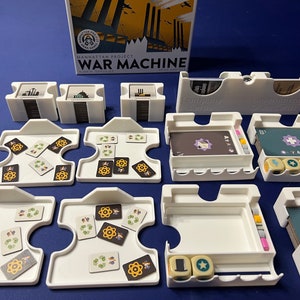 May include: A white plastic organizer for the board game Manhattan Project: War Machine. The organizer has compartments for cards, dice, and tokens. The game box is visible in the background.