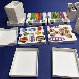 May include: A white 3D-printed board game organizer set featuring multiple modular trays, card holders, and token containers. The components are designed to hold various game pieces, including hexagonal tiles, colored cubes, and decks of cards.