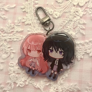Op de afbeelding: Een heldere acryl sleutelhanger met twee anime-achtige karakters. Eén karakter heeft roze haar en de ander heeft zwart haar. Beide karakters dragen outfits met rokken en jasjes. De sleutelhanger heeft een zilveren sluiting.