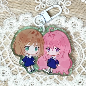May include: A clear acrylic keychain featuring two anime-style girls. One girl has brown hair and green eyes, the other has pink hair and purple eyes. Both are wearing blue dresses. The keychain has a white clasp.