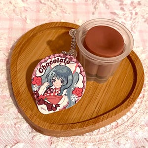 May include: A wooden heart-shaped tray holds a chocolate pudding cup and a keychain. The keychain features a cartoon girl with blue hair, the word "Chocolate", and snowflake accents. The pudding cup is brown.