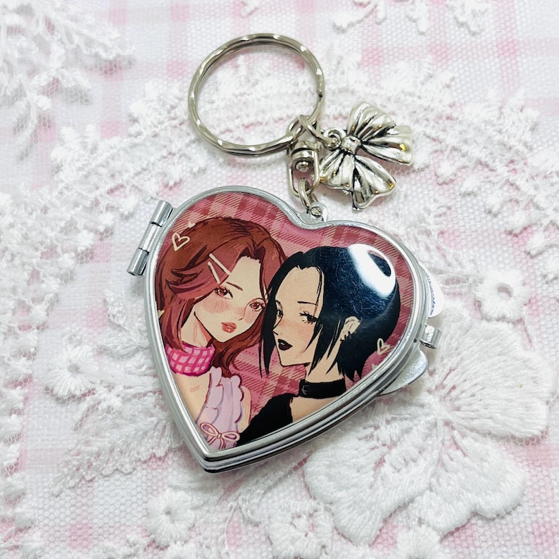May include: Heart-shaped silver keychain with a hinged locket. The locket features an anime-style illustration of two women. The keychain has a bow charm and a silver key ring. The background is a pink and white lace pattern.