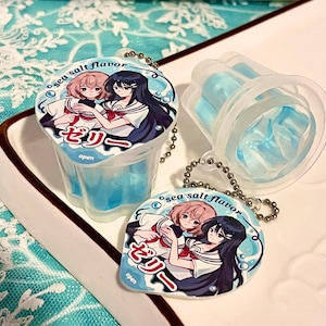 May include: Clear plastic cup filled with blue jelly, with a matching keychain. The keychain and cup lid feature an anime-style illustration of two girls in sailor uniforms. The text on the lid reads "sea salt flavor" and Japanese characters.