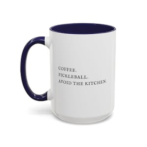 Peut inclure: Un mug en céramique blanche avec un intérieur et une anse bleu foncé. L'extérieur présente un texte noir qui dit : COFFEE. PICKLEBALL. AVOID THE KITCHEN. Le mug a un corps cylindrique avec une anse incurvée sur le côté gauche et une finition lisse.