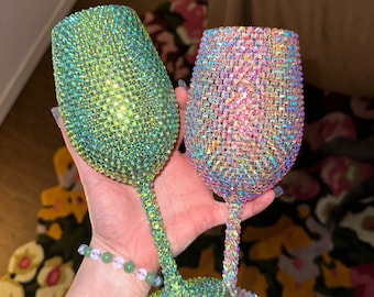 May include: Two iridescent wine glasses encrusted with sparkling rhinestones. One glass has a green hue, the other a pinkish-purple. The stems and bases are also covered in rhinestones, creating a glamorous look.