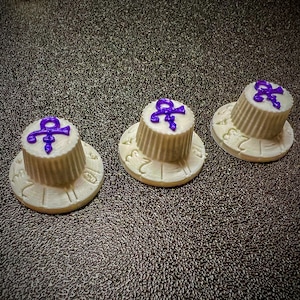 May include: Three white guitar knobs with a purple Prince symbol on top. The knobs have a textured, ribbed design and numbers around the edge.