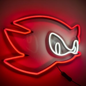 May include: A neon sign in the shape of a cartoon character's head, with a red outline and white details. The sign is mounted on a wall and has a black power cord.