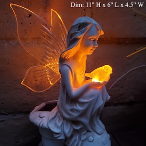 Può includere: Una statuetta di fata bianca con ali e un uccello nelle sue mani. La fata è seduta su una base a forma di fungo. La statuetta è illuminata da luci LED bianco caldo. Dim: 11" H x 6" L x 4.5" W