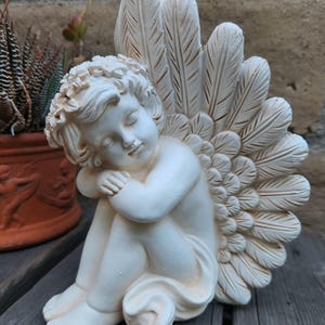 May include: A cream-colored cherub statue with closed eyes, seated and hugging its knees. The statue has large feathered wings and a floral crown. The cherub is on a wooden surface, with a potted plant in the background.