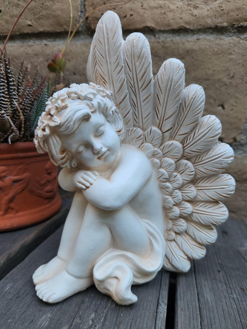 May include: A cream-colored cherub statue with closed eyes, seated and hugging its knees. The statue has large feathered wings and a floral crown. The cherub is on a wooden surface, with a potted plant in the background.