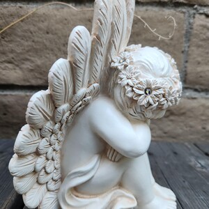 May include: A cream-colored angel figurine, seated with its knees drawn up. The angel has large, detailed feathered wings and a floral crown. The figurine is made of a smooth material and is placed on a wooden surface.