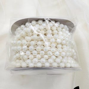 May include: A close-up of a spool of white faux pearl plastic beads, 6mm in size. The beads are strung on a clear thread and are packaged in a clear plastic bag. The text "6mm - White AB Faux Pearl Plastic Beads, 12 Yards" is visible on the packaging.