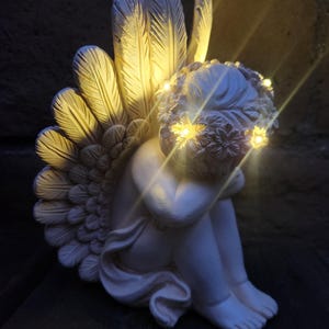May include: A white cherub statue with illuminated wings and a floral crown. The angel is seated, with its knees drawn up and arms wrapped around them. The wings are detailed with feather textures, and the crown and wings have small, warm-toned lights.
