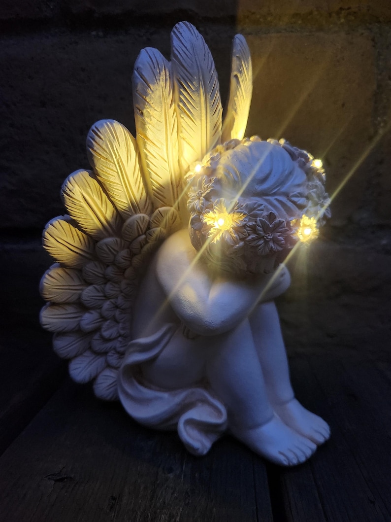May include: A white cherub statue with illuminated wings and a floral crown. The angel is seated, with its knees drawn up and arms wrapped around them. The wings are detailed with feather textures, and the crown and wings have small, warm-toned lights.