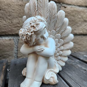 May include: A cream-colored cherub statue with a floral crown and feathered wings. The angel is seated with its arms wrapped around its knees. The statue is made of a smooth material and has a serene expression. The wings are detailed with individual feather markings.