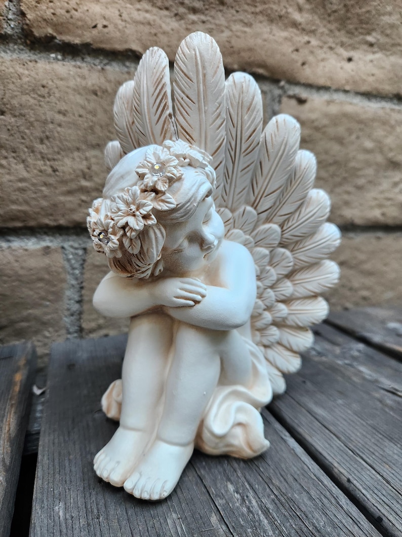 May include: A cream-colored cherub statue with a floral crown and feathered wings. The angel is seated with its arms wrapped around its knees. The statue is made of a smooth material and has a serene expression. The wings are detailed with individual feather markings.