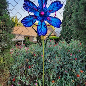 Könnte beinhalten: Ein Gartenstecker aus Metall mit einer großen, mehrschichtigen blauen Blume mit spitzen Blütenblättern und einer leuchtend rosa Mitte. Die Blume ist auf einem dünnen, grünen Metallstab befestigt. Das Design enthält geschwungene violette Akzente.