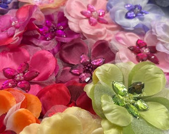 May include: A collection of multi-colored fabric flowers featuring layered petals and centers embellished with faceted crystals and small beads. The assortment includes shades of pink, purple, yellow, green, orange, and white in various sizes.