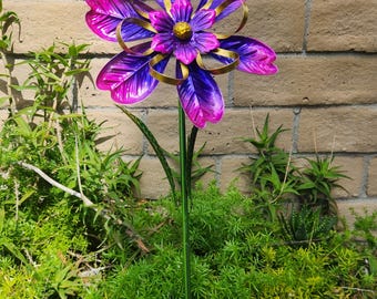 Könnte beinhalten: Ein Gartenstecker aus Metall mit einer großen, mehrschichtigen Blume mit leuchtend violetten und magentafarbenen Blütenblättern. Die Blume hat goldfarbene, geschwungene Akzente und eine gelbe, texturierte Kugel in der Mitte, montiert auf einem dünnen grünen Metallstab.