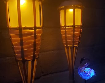 Könnte beinhalten: Zwei braune Korb-Tiki-Fackeln mit gelbem Licht und ein blauer Glasflamingo mit einem lila und blauen Muster und einem weißen Licht.